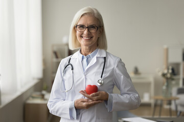 Happy senior doctor woman holding red heart shape object at chest, looking at camera with toothy smile. Positive mature practitioner, cardiologist in white uniform and glasses portrait