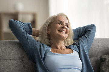 Cheerful retired elderly woman resting on home couch with soft back, stretching arms, open elbows,...
