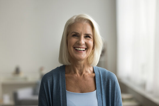 Happy blonde senior retired woman looking at camera with perfect toothy smile, posing at home, laughing. Cheerful positive pretty older lady head shot video call front portrait
