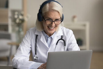Positive senior doctor woman in wireless headphones speaking on online video call to patient,...