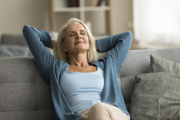 Peaceful satisfied elderly woman relaxing on couch with closed eyes, smiling, stretching body,...