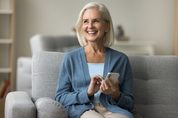 Cheerful pretty senior woman relaxing on sofa with digital device, holding smartphone, using online application for Internet communication, looking away, thinking, smiling, laughing