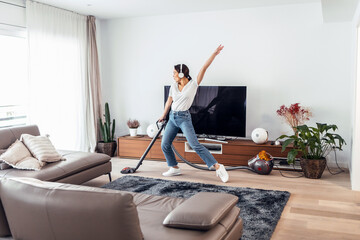 Young happy woman listening and dancing to music while cleaning the living room floor with a vaccum...