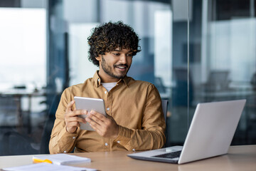 Young successful hispanic man with tablet computer, satisfied with achievement results smiling looking at screen, man using application, sitting at workplace with laptop inside office.