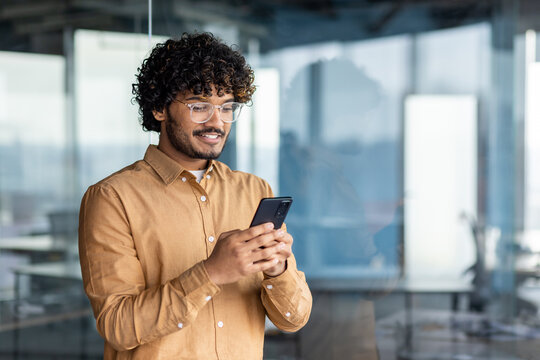 Hispanic Man Smiles And Looks At The Phone Screen, Businessman Inside The Office Holds A Smartphone, Browses Online Internet Pages, Makes A Call, And Uses An Application, Near The Window In A Shirt.