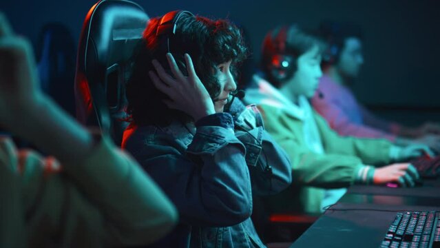 Medium shot of young multiracial girl with curly hair, in denim jacket sitting in chair at cyber cafe, putting on headphones, preparing to play first person shooter multiplayer game