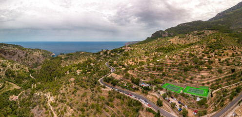 The panoramic view of Deia at the Island Mallorca shows the treelined mountain road from a high point near the ocean, Mountain and Bay on the mediterranean coastline, Tennis court and cloudy sky