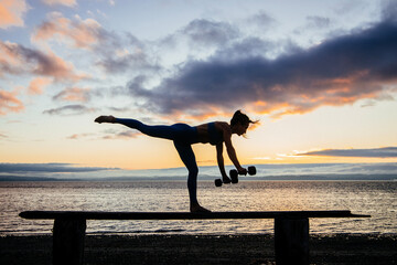 Silhouette of young woman doing workout with weights on a bench in front of ocean sunrise or sunset