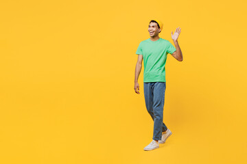 Fototapeta na wymiar Full body smiling fun happy cheerful young man of African American ethnicity he wearing casual clothes green t-shirt hat walking going waving hand isolated on plain yellow background studio portrait.