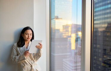 Young Asian business woman professional executive manager, company employee using digital tablet fintech looking at smart pad in corporate office standing at big window, city view. Copy space