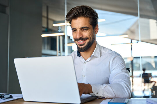 Happy Young Latin Business Man Looking At Laptop At Work. Smiling Businessman Professional Employee, Company Manager Using Computer Sitting At Desk Managing Digital Data In Office.