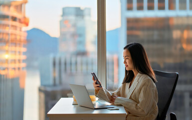Young busy Asian business woman leader holding cellphone device using mobile phone, looking at smartphone checking financial market app working in modern corporate office with big window. - Powered by Adobe