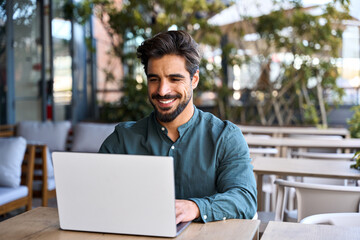 Happy young Latin business man using laptop sitting outdoors. Smiling Hispanic guy student or...