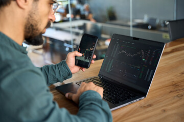 Investor using mobile phone and laptop checking trade market data. Stock trader broker looking at...