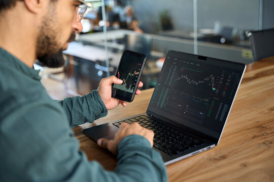 Investor using mobile phone and laptop checking trade market data. Stock trader broker looking at computer analyzing trading cryptocurrency finance market crypto stockmarket data, over shoulder view.