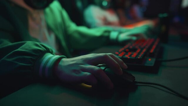 Close-up shot of hands of anonymous young guy in green casual bomber jacket manipulating computer mouse and using keyboard while playing shooter game in dark cyber cafe