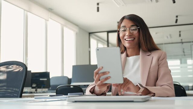 Young happy smiling professional African American business woman manager sales executive wearing suit and glasses, using digital tablet working sitting at desk in office looking at tab.