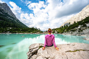 Woman sits on big rock at turquoise Sorapis lake and enjoys the view on impressive Cadini di Misurina in the afternoon. Lake Sorapis, Dolomites, Belluno, Italy, Europe.