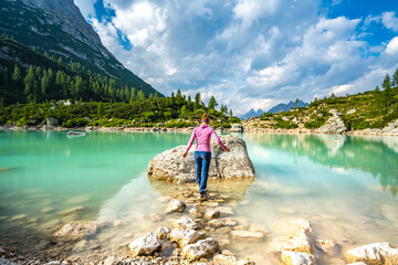 Fototapeta na wymiar Athletic young woman hops over stone path to large rock at beautiful turquoise Sorapis lake in the afternoon. Lake Sorapis, Dolomites, Belluno, Italy, Europe.