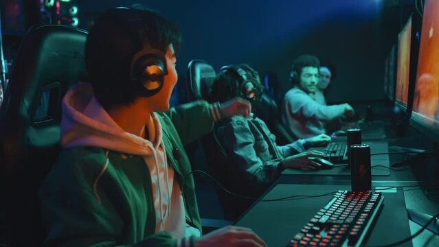 Medium side shot of young Chinese gamer in headset and multinational team mates playing FPS game tournament, winning, cheering, whooping and celebrating with high fives