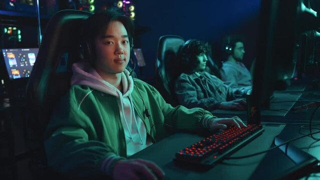 Medium portrait shot of young Chinese man in green bomber jacket sitting in cyberclub playing video games with friends, turning head and looking at camera