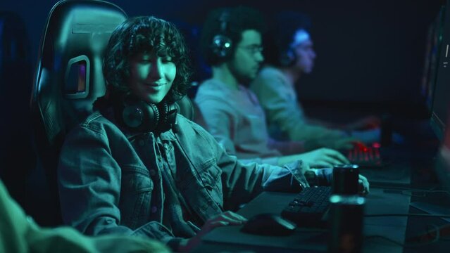 Medium portrait shot of group of diverse people sitting in cyberclub playing video games, and young multiracial woman with curly hair, in denim jacket taking off headphones and smiling at camera