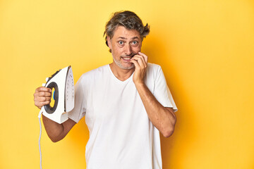 Man with iron in hand on a yellow background biting fingernails, nervous and very anxious.