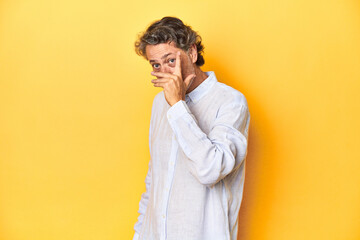 Middle-aged man posing on a yellow backdrop blink at the camera through fingers, embarrassed covering face.