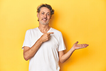 Middle-aged man posing on a yellow backdrop excited holding a copy space on palm.