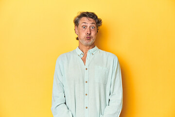 Middle-aged man posing on a yellow backdrop blows cheeks, has tired expression. Facial expression concept.