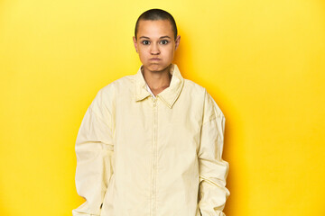 Young woman in jacket, yellow studio backdrop blows cheeks, has tired expression. Facial expression...
