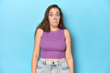 Fashionable young woman in a purple top on blue background shrugs shoulders and open eyes confused.