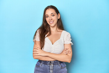 Stylish young woman in white blouse on a blue studio backdrop who feels confident, crossing arms with determination.