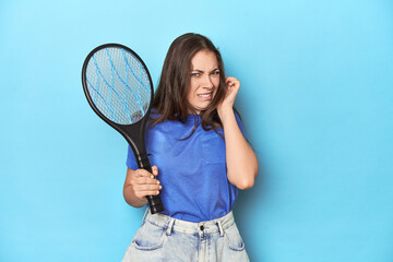Woman with an electric bug zapper on a blue background covering ears with hands.
