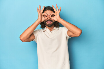 Long-haired man in a white polo, blue studio showing okay sign over eyes