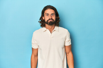 Long-haired man in a white polo, blue studio shrugs shoulders and open eyes confused.