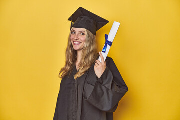 Young caucasian woman wearing a graduation robe holding a diploma Ylooks aside smiling, cheerful...