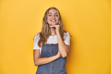 Young blonde Caucasian woman in denim overalls posing on a yellow background, smiling happy and confident, touching chin with hand. - obrazy, fototapety, plakaty