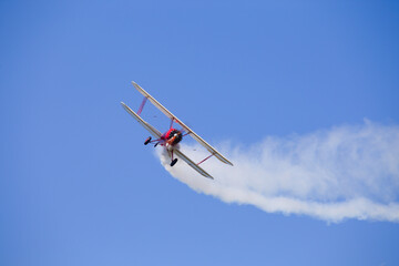 A red biplane Hatz Classic with white wings and radial engine blows white smoke behind and turns against a blue cloudless sky.