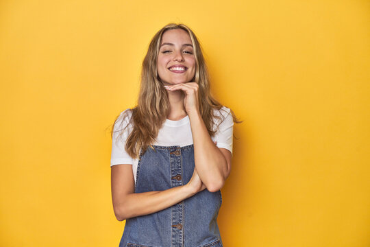 Young blonde Caucasian woman in denim overalls posing on a yellow background, smiling happy and confident, touching chin with hand.