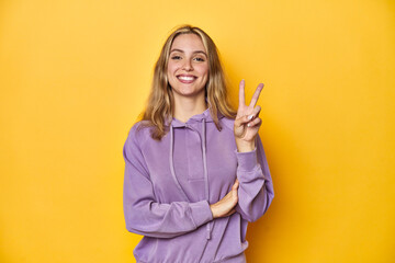 Young blonde Caucasian woman in a violet sweatshirt on a yellow background, showing number two with...