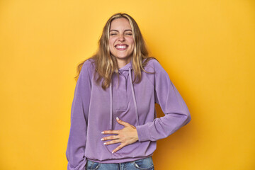 Young blonde Caucasian woman in a violet sweatshirt on a yellow background, touches tummy, smiles...