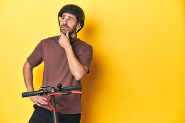 Man with electric scooter and helmet, yellow studio looking sideways with doubtful and skeptical expression. © Asier