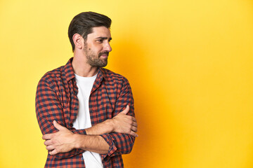 Caucasian man in red checkered shirt, yellow backdrop smiling confident with crossed arms.