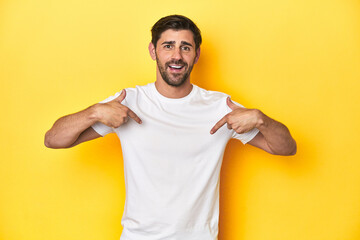 Caucasian man in white t-shirt on yellow studio background surprised pointing with finger, smiling...