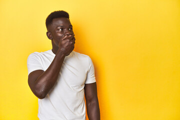 Stylish young African man on vibrant yellow studio background, thoughtful looking to a copy space covering mouth with hand.