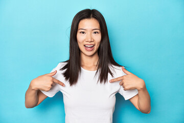 Young Asian woman in white t-shirt, studio shot, surprised pointing with finger, smiling broadly.