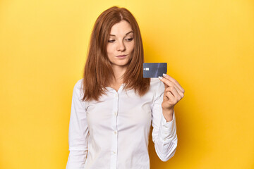 Redhead holding credit card, financial concept confused, feels doubtful and unsure.