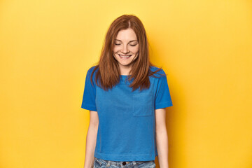 Redhead in blue t-shirt on yellow backdrop laughs and closes eyes, feels relaxed and happy.