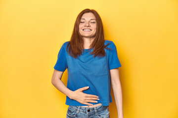 Redhead in blue t-shirt on yellow backdrop touches tummy, smiles gently, eating and satisfaction...